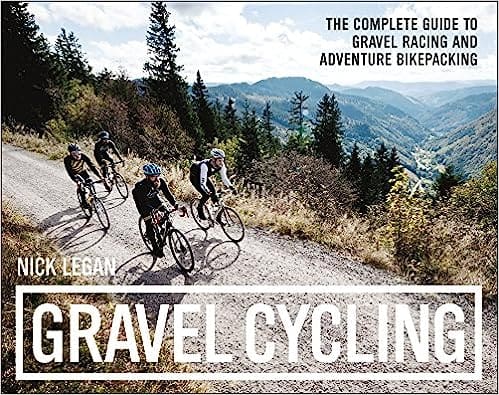 Three cyclists skillfully navigating a sharp turn on a high mountain dirt road, with a panoramic view of distant mountains, tree lines, and clear sky in the background.