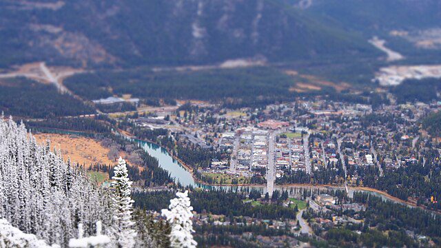 Aerial view of Banff townsite in autumn with snow-dusted evergreen trees in foreground and mountains in background, showing typical banff weather in october conditions with mixed snow and fall colors