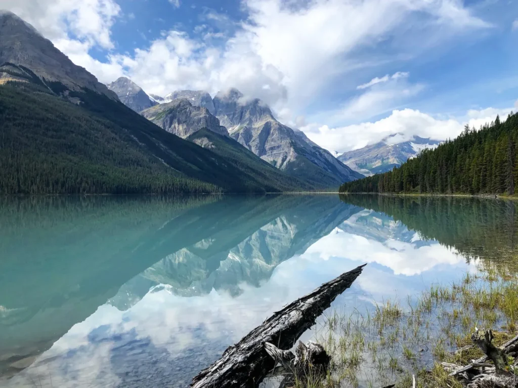 Glacier Lake Banff with calm turquoise water reflecting the surrounding mountains and forest under a bright sky.