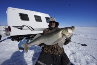 Ice angler holds large walleye fish caught using best line for ice fishing walleye, standing on Lake Winnipeg with ice fishing shelter in background.