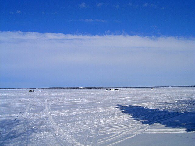 Ice fishing in Alberta at Pigeon Lake with snowmobile tracks, fishing shelters, and vehicles scattered across the frozen lake surface under a bright blue sky with wispy clouds.